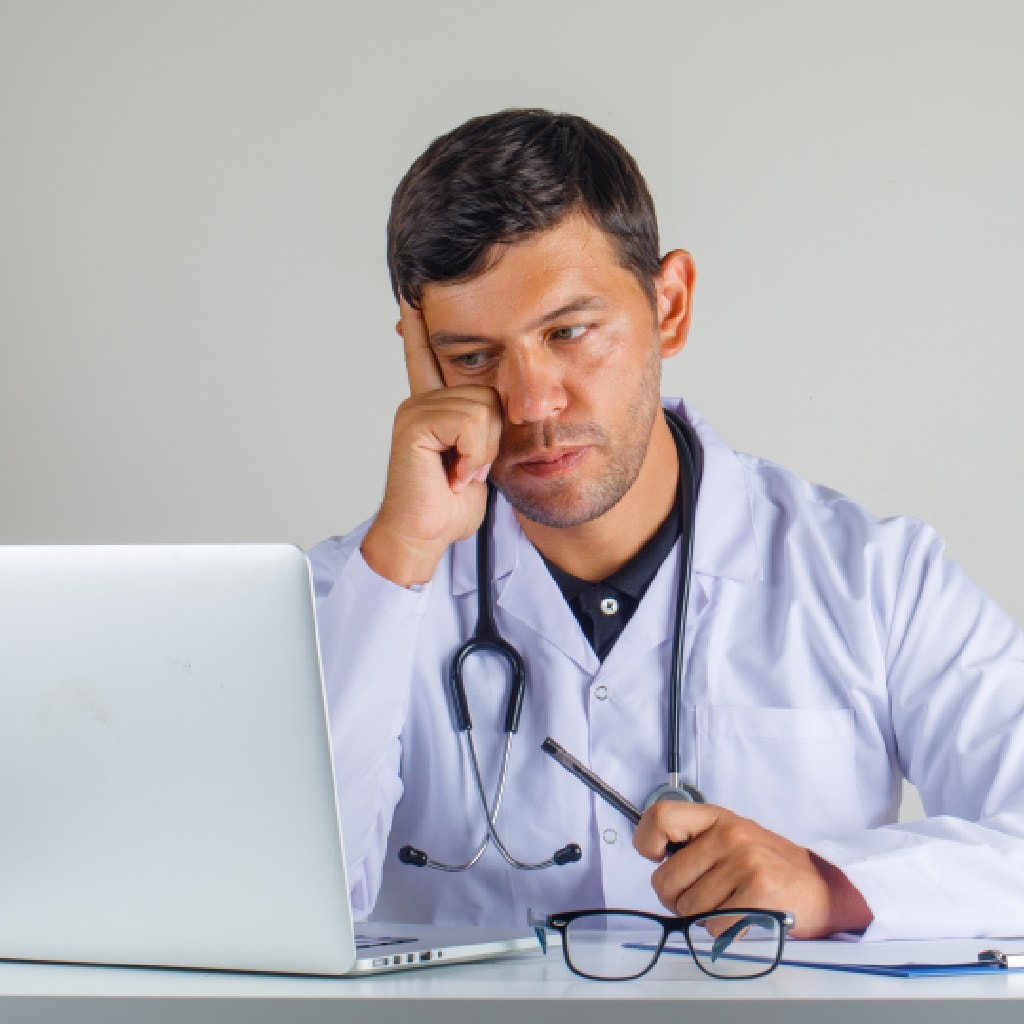 A thoughtful doctor in a white coat sits at a desk, staring intently at a laptop. A stethoscope hangs around his neck, with eyeglasses and a clipboard nearby.