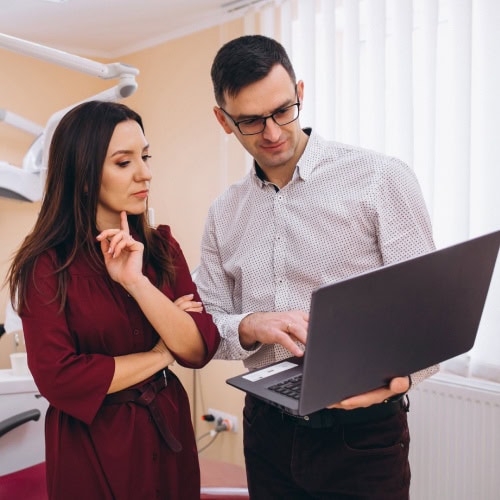 A woman in a burgundy dress and a man in glasses and a white shirt are looking at a laptop in a bright office. They appear focused and engaged.