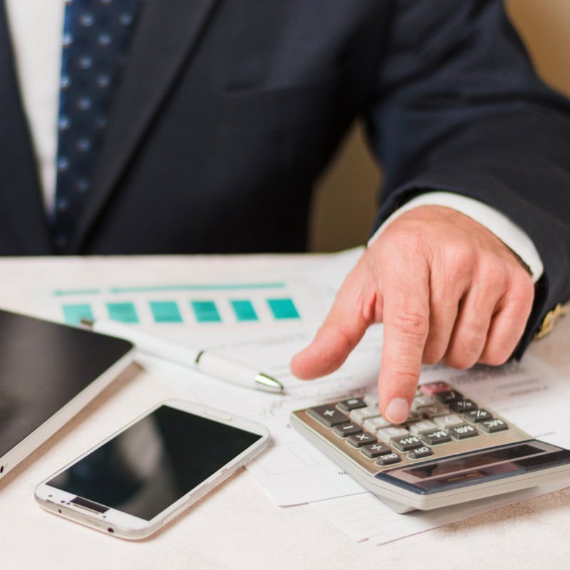 A person in a suit uses a calculator on a table with charts, a smartphone, and a tablet; conveying a business and analytical setting.