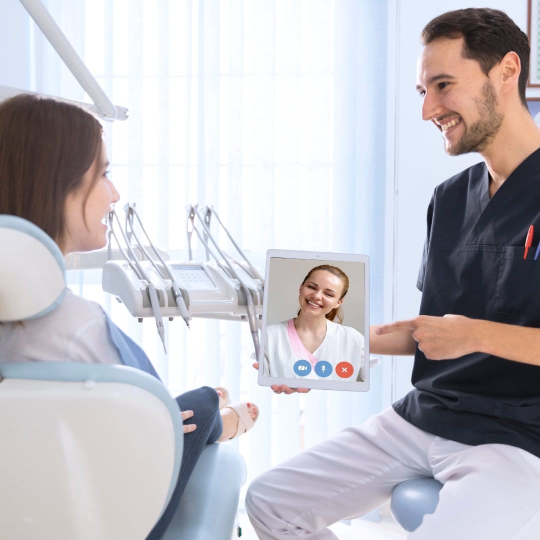 A dentist holds a tablet showing a smiling woman on a video call, chatting with a patient seated in a dental chair. The scene is bright and friendly.