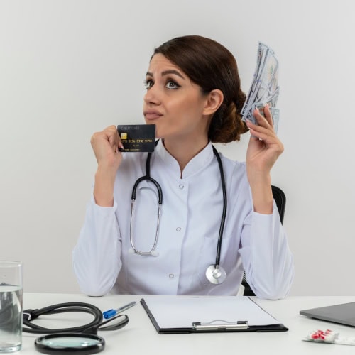A female doctor in a white coat holds cash and a credit card, appearing thoughtful. A stethoscope, clipboard, pills, and water are on the desk.