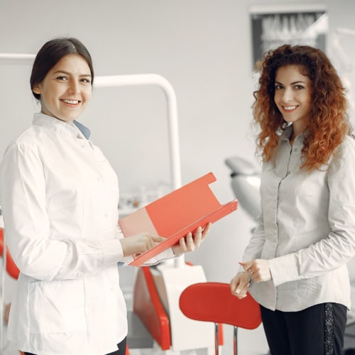 Two women in a dental office smile at the camera. One wears a white coat and holds a red folder, suggesting a professional, friendly environment.