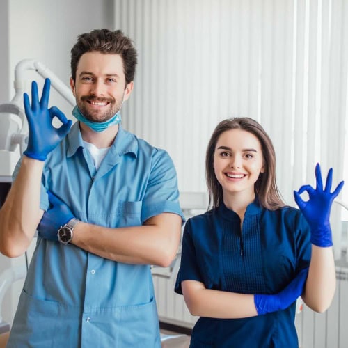 Two smiling dental professionals in blue scrubs and gloves make OK signs, standing in a dental office, conveying a reassuring and positive atmosphere.