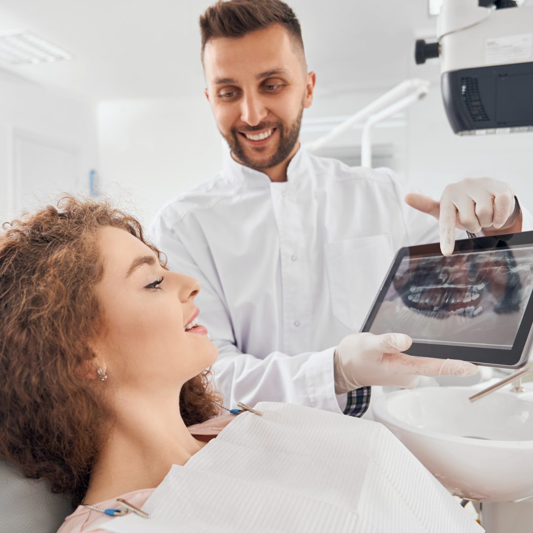 A dentist shows a smiling female patient her dental X-ray on a tablet. The setting is a bright, modern dental office, suggesting a positive atmosphere.
