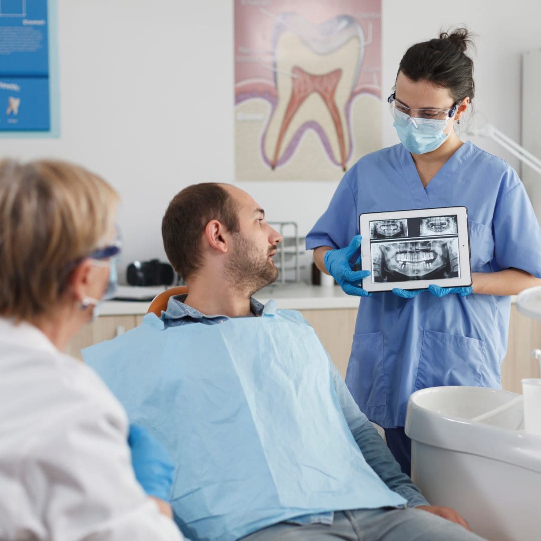 Dentist in blue scrubs shows a patient dental X-rays on a tablet. The patient, wearing a bib, looks at the screen, while another staff member observes.