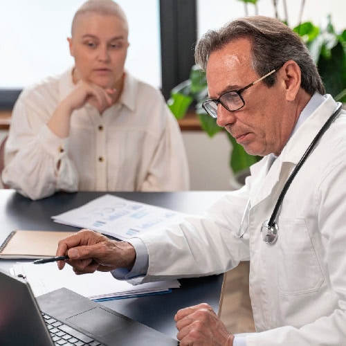 A doctor in a white coat points to a laptop screen, discussing something with a patient. The patient listens attentively, creating a focused atmosphere.