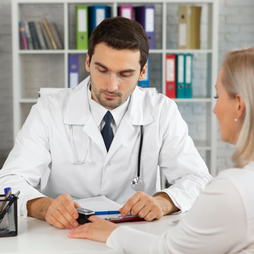 A doctor in a white coat attentively listens to a patient in an office setting, with shelves of colorful binders in the background.