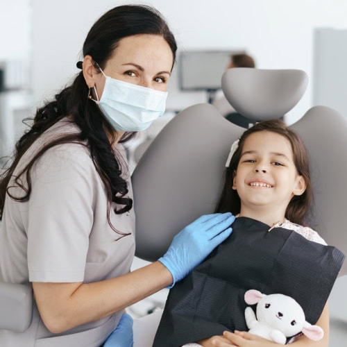 Dentist wearing a mask and gloves smiles beside a young girl in a dental chair holding a stuffed toy. The setting is bright and welcoming.